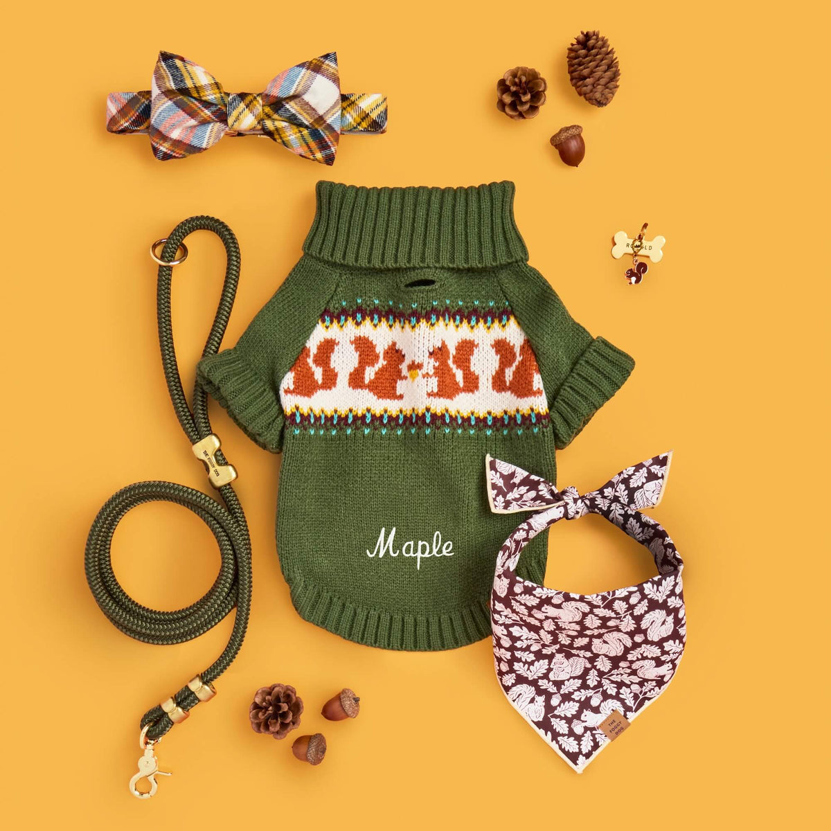 A styled flat-lay of a green knit pet sweater with a white and orange Fair Isle pattern across the chest, personalized with the name 'Maple' in white script. The sweater is displayed with a matching green leash and accessories including a plaid bow tie, patterned bandana, and small decorative items on a solid orange background.