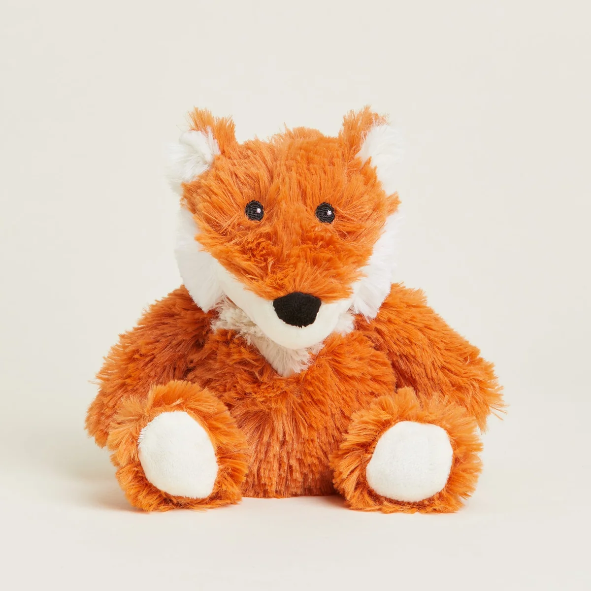 A plush toy fox made of soft orange-brown faux fur with white accents on its muzzle, inner ears, and paws, sitting against a plain light beige background.