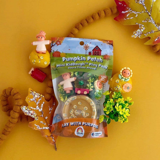 Pumpkin Patch Mini KidDough Play Pack toy packaging shown on a bright yellow background with autumn-themed decorative props including a small pumpkin figure, glittery leaves, and small plants.