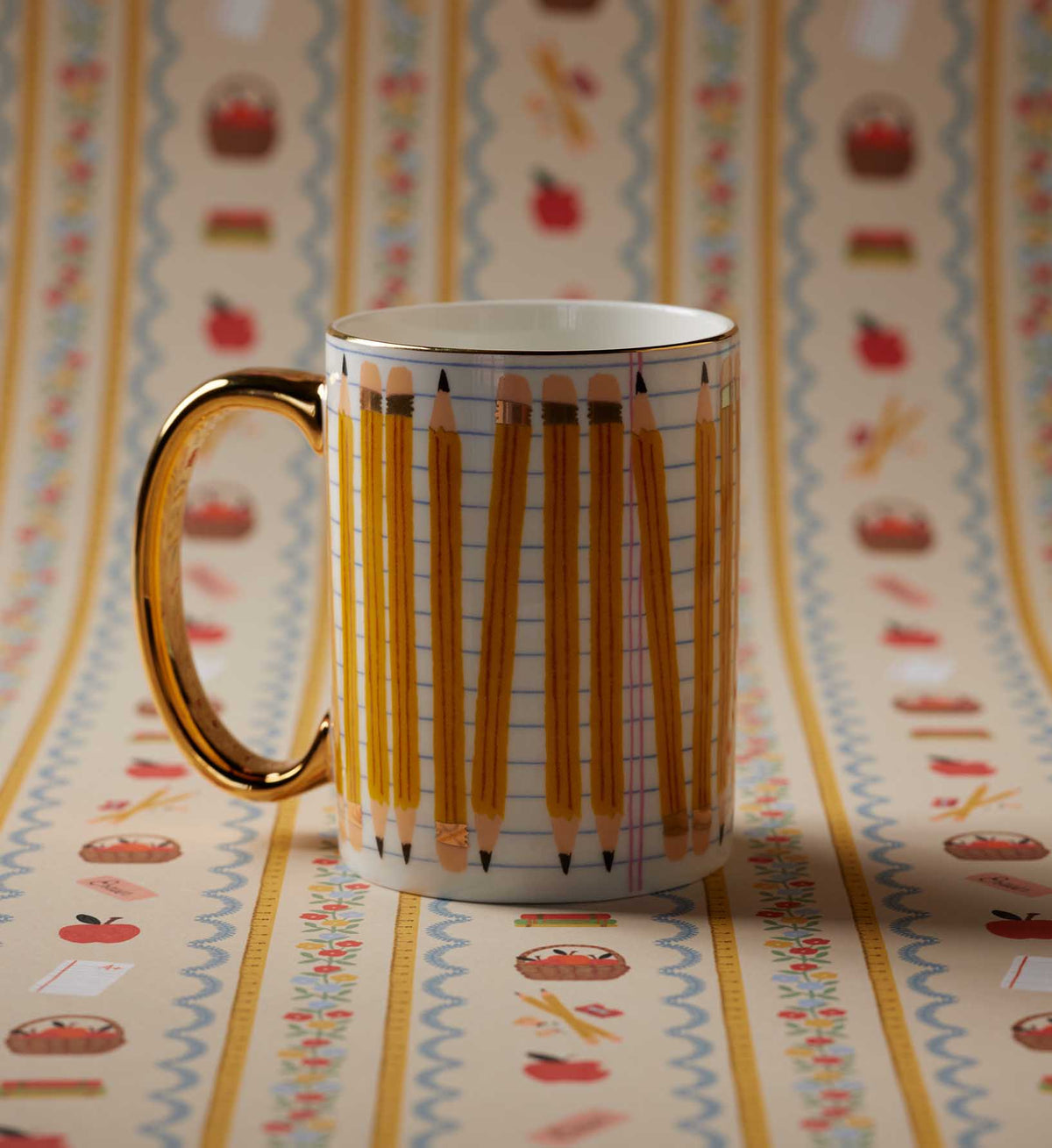 A white ceramic mug with a gold handle and gold rim, decorated with a pattern of yellow pencils on graph paper, set against a matching patterned background.